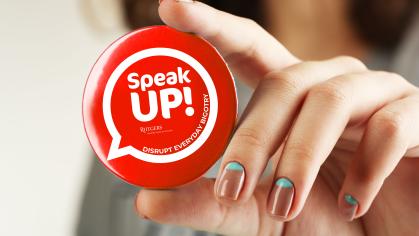 A hand holding a round button that says Speak up, disrupt everyday bigotry