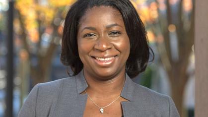 Anna Branch wearing a grey jacket and black and white blouse standing in front of trees during the fall on Rutgers campus 