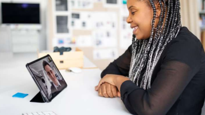 An african american woman with grey and white braids sitting in a classroom speaking to a mentor on a tablet