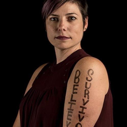 Laura Luciano, Associate Director for the VPVA Office on the Camden Campus, looking forward, focused, wearing a sleeveless black blouse, against a black background, &quot;Believe Survivors&quot; is written in black on her arm.