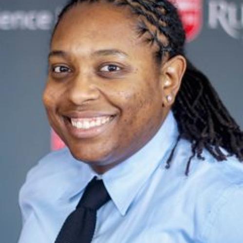 Avery Arrington - an african american woman smiling against a Rutgers posters. Her hair is in locks and she is wearing a light blue shirt and dark blue tie