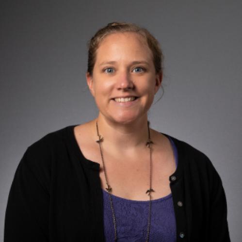 Christina Bifulco is smiling against a grey background. She a caucasian woman with her hair slicked back into a bun. She is wearing a purple inside shirt and a black cardigan