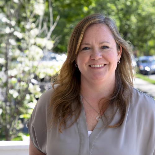 Kerri Wilson - closeup of caucasian woman with blue eyes and blond highlights. She is standing on a porch and smiling while wearing a light grey banded collar shirt.