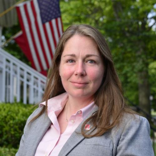 Ann Treadaway is a caucasian woman with long nrown and gray hair. She is smiling infront of an american flag on a house. She is weraing a pink collared shirt and a grey jacket