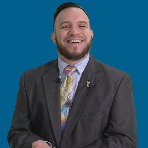 Gary Mendoza is a spanish mal. He is standing angainst a blue background and he is smiling. He is wearing a dark grey suit with a light grey collared shirt,a yellow tie with a necklace on the tie.