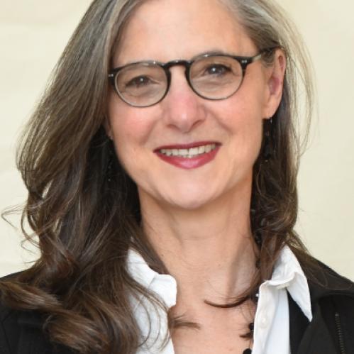Nancy is a jewish woman with long grey and black hair. She is smiling and wearing black rimmed glasses and a white shirt with a black jacket