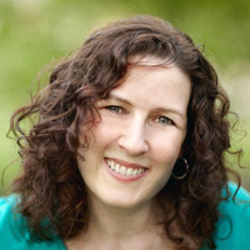 Karen Small - A jewish woman with curly dark brown hair. She is looking ahead and smiling and wearing a teal shirt while standing against a green plant background