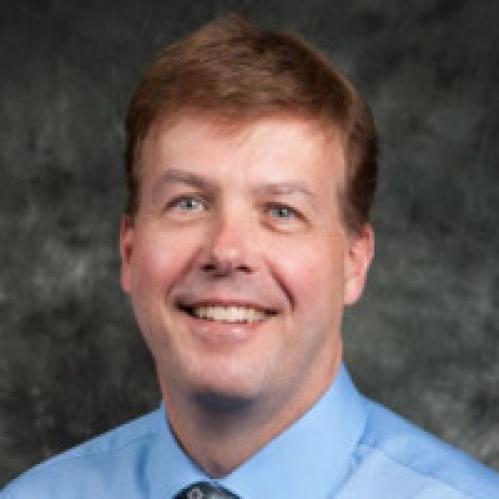 Bill Welsh is a caucasian man with short hair. He is wearing a blue collared shirt ad standing in a classroom.