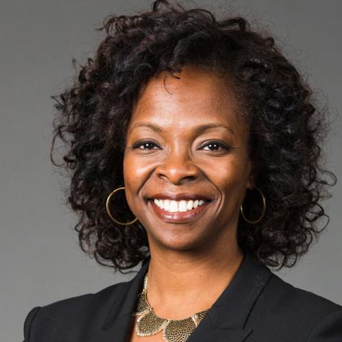 Corlisse Thomas is an african american woman with curly shoulder length hair. She is smiling and standing in front of a grey wall and wearing a black suit jacket with a gold necklace.
