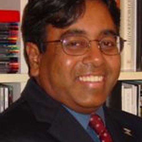 Edward is an indian man who is smiling a in front of a book shelf. He is wearing a blue shirt and a red polka dot tie and a black jacket..