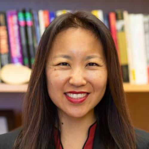 Ji Lee is an asian american woman with long brown hair. She is wearing red lipstick and smiling while standing in front of a book shelf. She is wearing a red collared shirt under a black jacket.