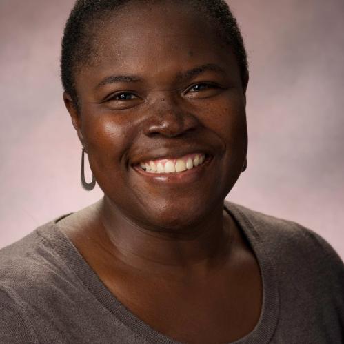 Kimberly is an african american woman with short hair. She is smiling and wearing a grey blouse and standing in front of a brown background