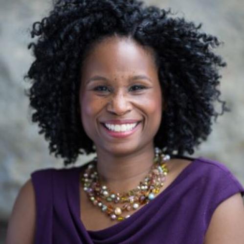 Nyeema is an african american woman with freckles and curly hair. She us smiling and wearing a purple blouse and a gold multicolored necklace.