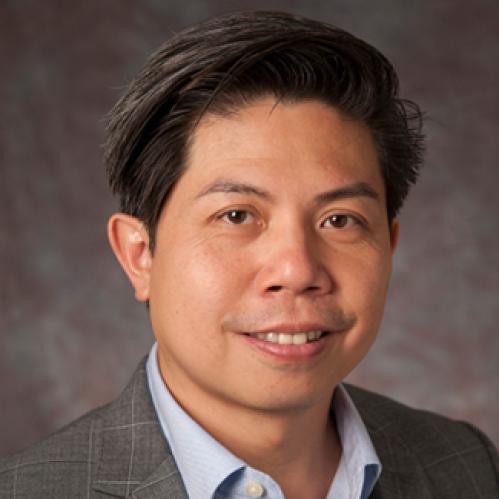 Rick is an asian man, smiling and he is wearing a light blue, collared shirt with a plaid grey jacket