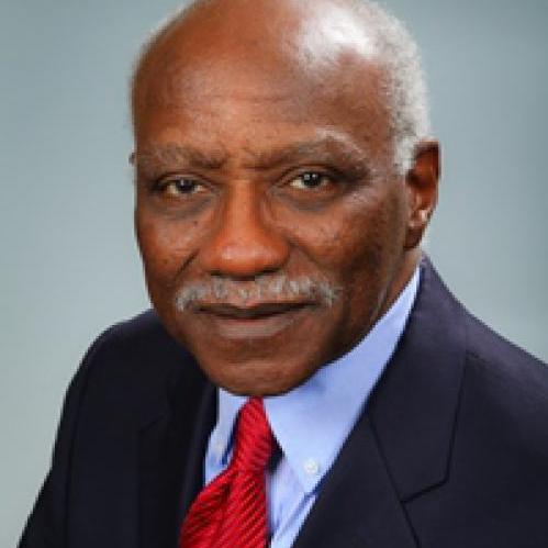 Ronald Quincy is an older african american man. He has grey hair and he is smiling and wearing a blue collared shirt, a red tie and a black jacket