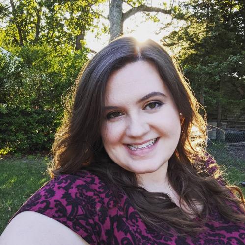Desiree has long brown hair and fair skin. She is smiling and wearing pink lipstick with a purple and black blouse. She is standing in a wooded area with the sunlight behind her head.