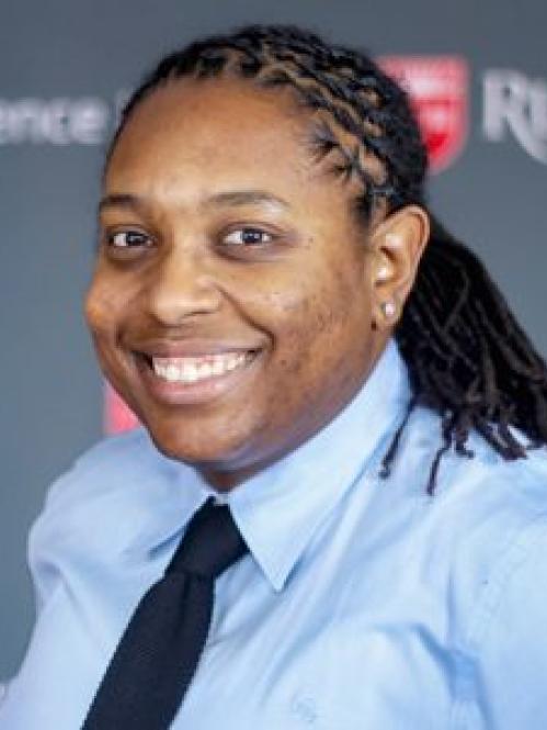 Avery Arrington - an african american woman smiling against a Rutgers posters. Her hair is in locks and she is wearing a light blue shirt and dark blue tie