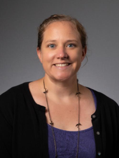 Christina Bifulco is smiling against a grey background. She a caucasian woman with her hair slicked back into a bun. She is wearing a purple inside shirt and a black cardigan