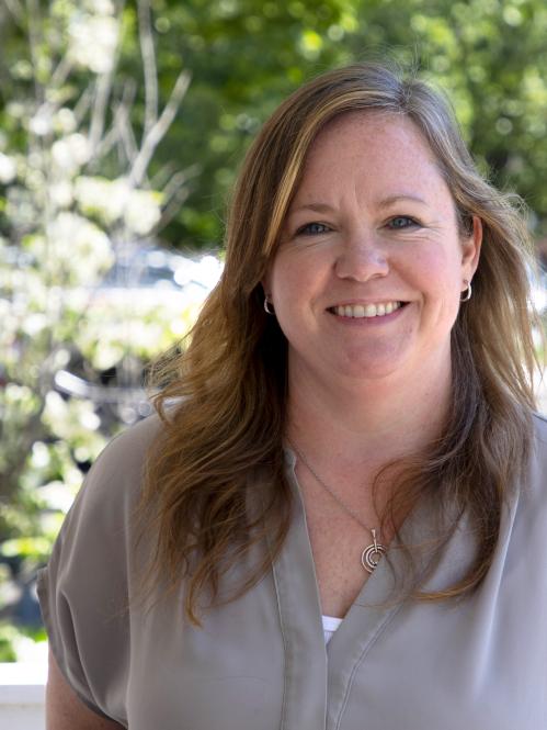 Kerri Wilson - closeup of caucasian woman with blue eyes and blond highlights. She is standing on a porch and smiling while wearing a light grey banded collar shirt.