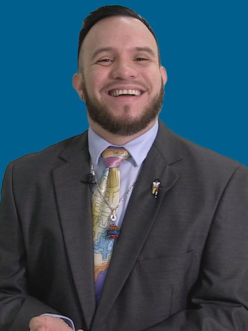 Gary Mendoza is a spanish mal. He is standing angainst a blue background and he is smiling. He is wearing a dark grey suit with a light grey collared shirt,a yellow tie with a necklace on the tie.