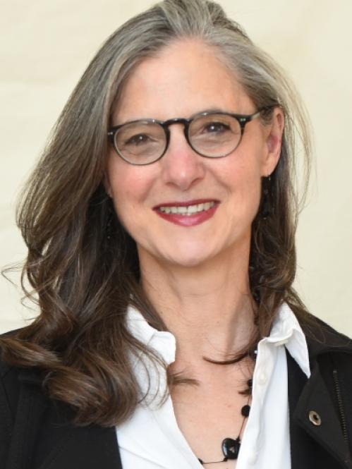 Nancy is a jewish woman with long grey and black hair. She is smiling and wearing black rimmed glasses and a white shirt with a black jacket