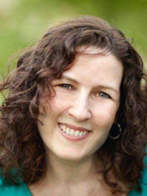 Karen Small - A jewish woman with curly dark brown hair. She is looking ahead and smiling and wearing a teal shirt while standing against a green plant background 