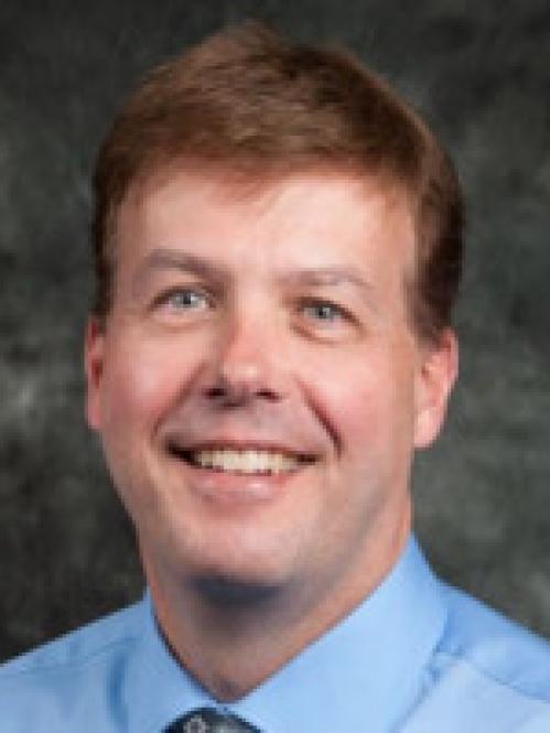 Bill Welsh is a caucasian man with short hair. He is wearing a blue collared shirt ad standing in a classroom.