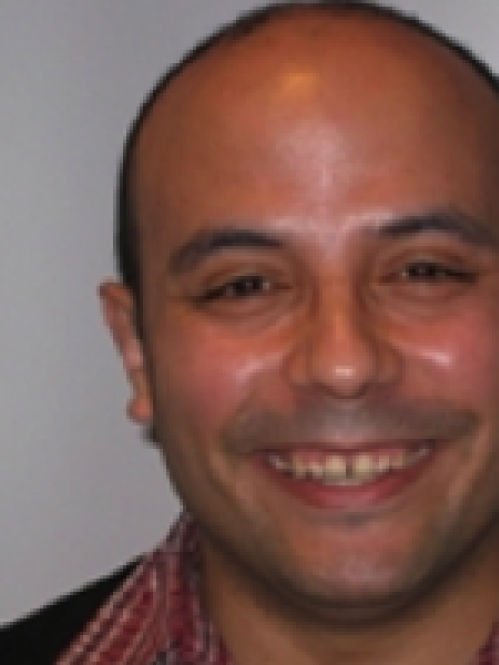 Carlos is a hispanic male with closely cut hair. He is smiling and wearing a red checkered collared shirt and a black jacket