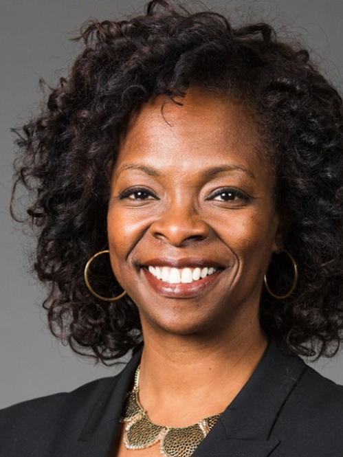 Corlisse Thomas is an african american woman with curly shoulder length hair. She is smiling and standing in front of a grey wall and wearing a black suit jacket with a gold necklace. 
