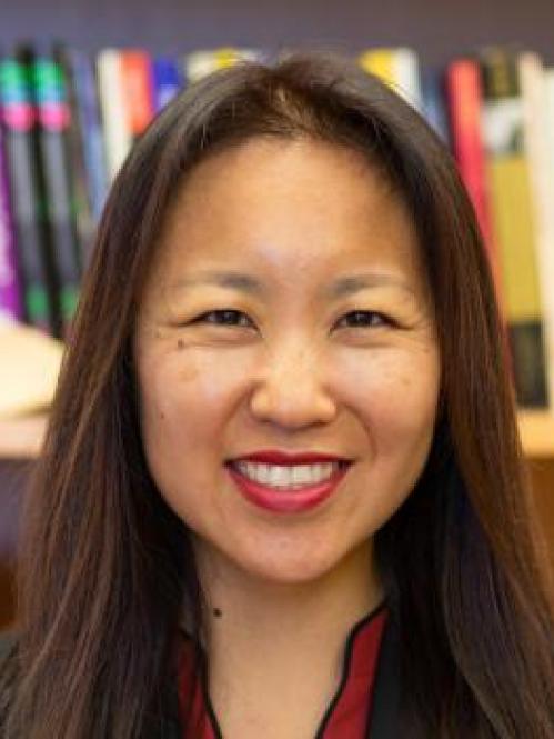 Ji Lee is an asian american woman with long brown hair. She is wearing red lipstick and smiling while standing in front of a book shelf. She is wearing a red collared shirt under a black jacket. 