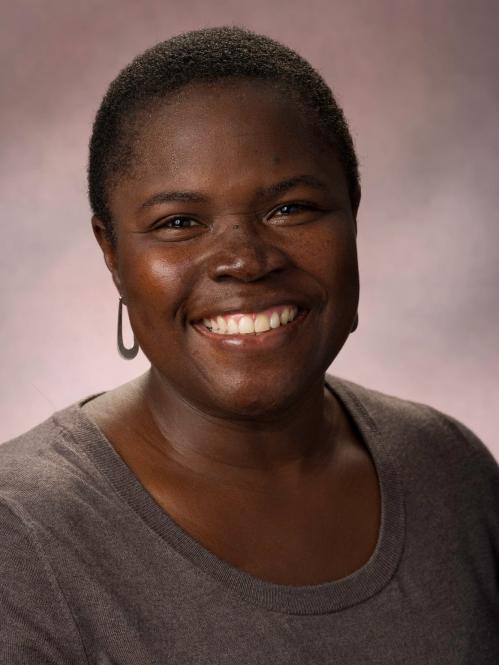 Kimberly is an african american woman with short hair. She is smiling and wearing a grey blouse and standing in front of a brown background