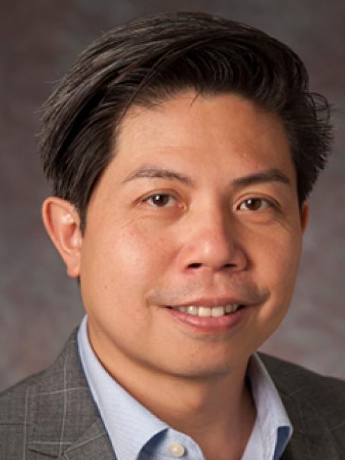 Rick is an asian man, smiling and he is wearing a light blue, collared shirt with a plaid grey jacket