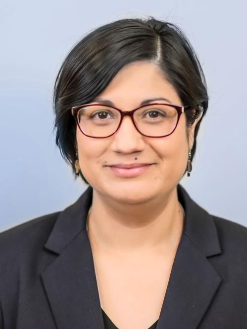 Corinne has short straight hair and olive skin. She is smiling and wearing red rimmed glasses and a black jacket and blouse