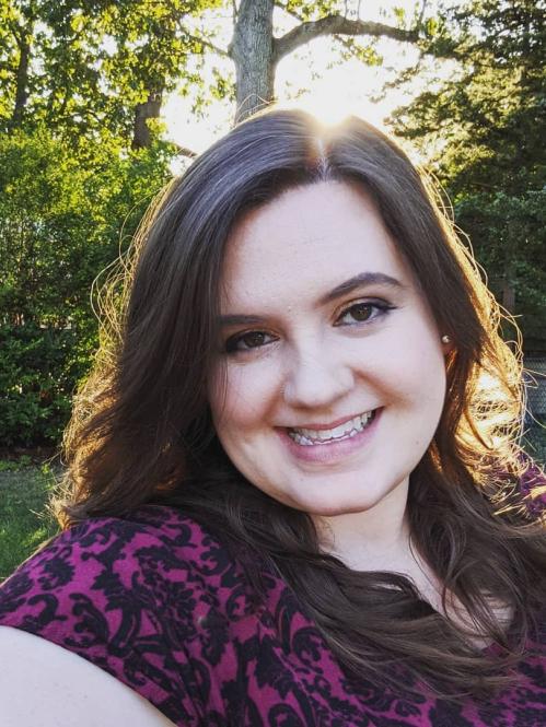 Desiree has long brown hair and fair skin. She is smiling and wearing pink lipstick with a purple and black blouse. She is standing in a wooded area with the sunlight behind her head.