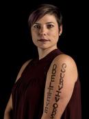 Laura Luciano, Associate Director for the VPVA Office on the Camden Campus, looking forward, focused, wearing a sleeveless black blouse, against a black background, "Believe Survivors" is written in black on her arm.