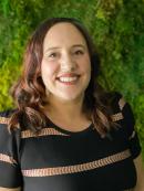 Crystal Bedley is smiling in front of a green background. she is wearing a black and brown striped dress
