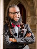 Keywaun is an african american man with well kept locs and beard. He is smiling and wearing a black suit and bow tie