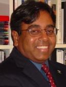 Edward is an indian man who is smiling a in front of a book shelf. He is wearing a blue shirt and a red polka dot tie and a black jacket.. 