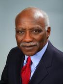 Ronald Quincy is an older african american man. He has grey hair and he is smiling and wearing a blue collared shirt, a red tie and a black jacket
