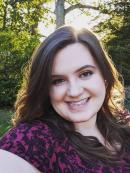 Desiree has long brown hair and fair skin. She is smiling and wearing pink lipstick with a purple and black blouse. She is standing in a wooded area with the sunlight behind her head.