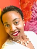 Joan Collier is an african american woman with short cropped hair. She is smiling and wearing red lipstick, a white pearl necklace and white dress. She is standing in front a rainbow painted wall and flower background.