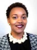 Joan Collier is an african american woman with short cropped hair. She is smiling and wearing red lipstick, a white pearl necklace and white dress. She is standing in front a rainbow painted wall and flower background.