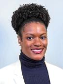 Joy is an african american woman. She is smiling and has curly hair with her her up in a bun. She is wearing a white jacket and a black turtle neck.