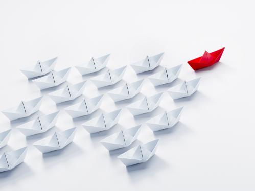 Paper boats on a white background moving in the shape of an arrow being lead by a red paper boat.
