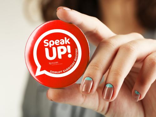 A hand holding a round button that says Speak up, disrupt everyday bigotry